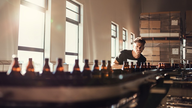 Man inspecting brown glass beer bottles on a conveyor belt in a modern brewery with sunlight and packaging boxes.