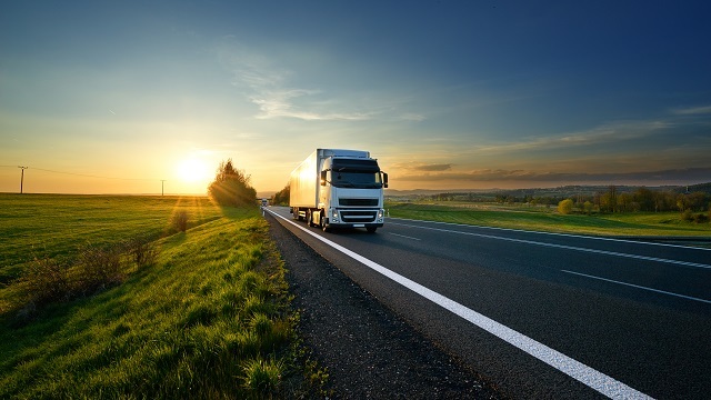 A white semi‑truck driving along a rural highway at sunset, surrounded by open fields and warm golden light, representing long‑haul transportation and logistics